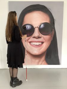Leona is standing to the side of and facing a six-foot canvas on which is painted a hyper-realistic close-up of her head. In the painting she's wearing purple and gold sunglasses (which sit slightly askew, and which reflect the lighting umbrellas) and smiling. In the studio she's standing in a black wrap dress, wearing black Dr Martens, and holding Moses, who bisects the painting from the sunglasses downward. Her black sunglasses are perched on her head. The painting is by Roy Nachum, and the photograph was taken in Roy’s studio Photo by Alabaster Rhumb.